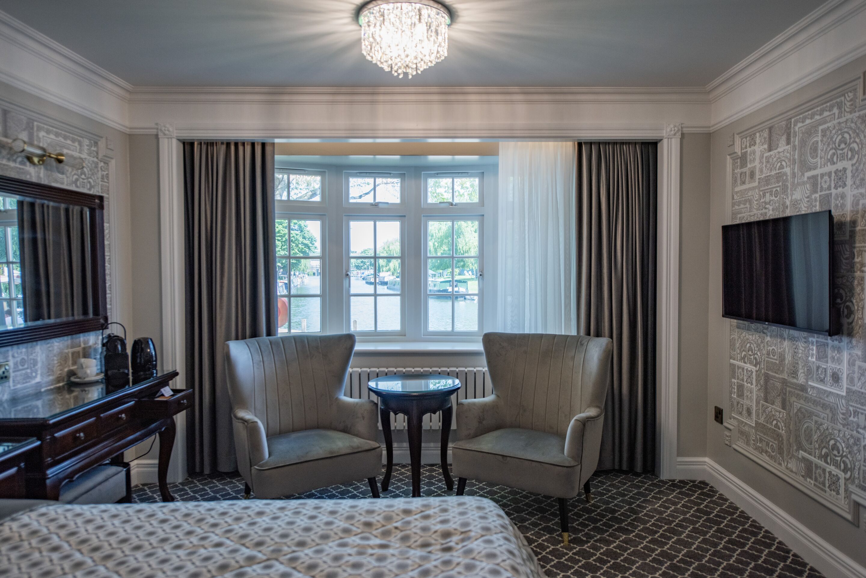 A hotel room with a bed, two armchairs, a small table, a television, and a large window with curtains.