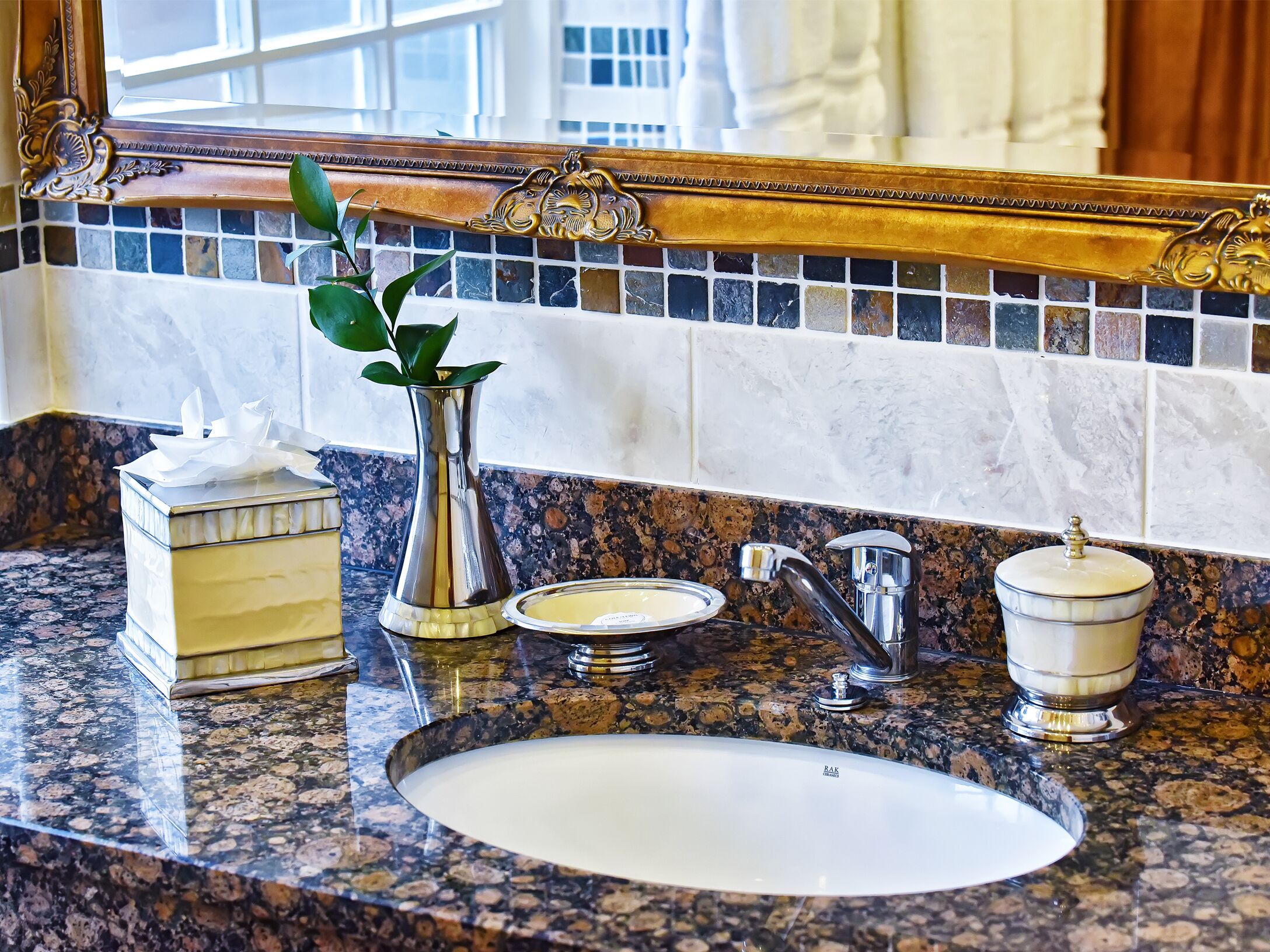 A bathroom with a granite countertop, a white sink, a mirror with ornate gold detailing, a vase with a plant, a tissue box, and a soap dish.