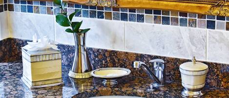 A bathroom with a granite countertop, a white sink, a mirror with ornate gold detailing, a vase with a plant, a tissue box, and a soap dish.