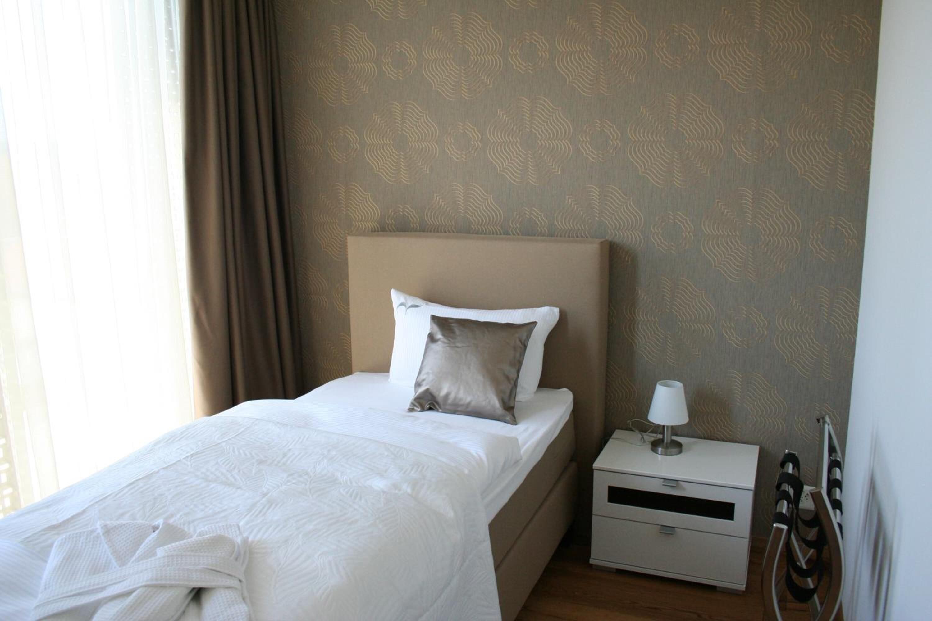 A neatly made bed with a white comforter and two pillows, a bedside table with a lamp, and a patterned wall.