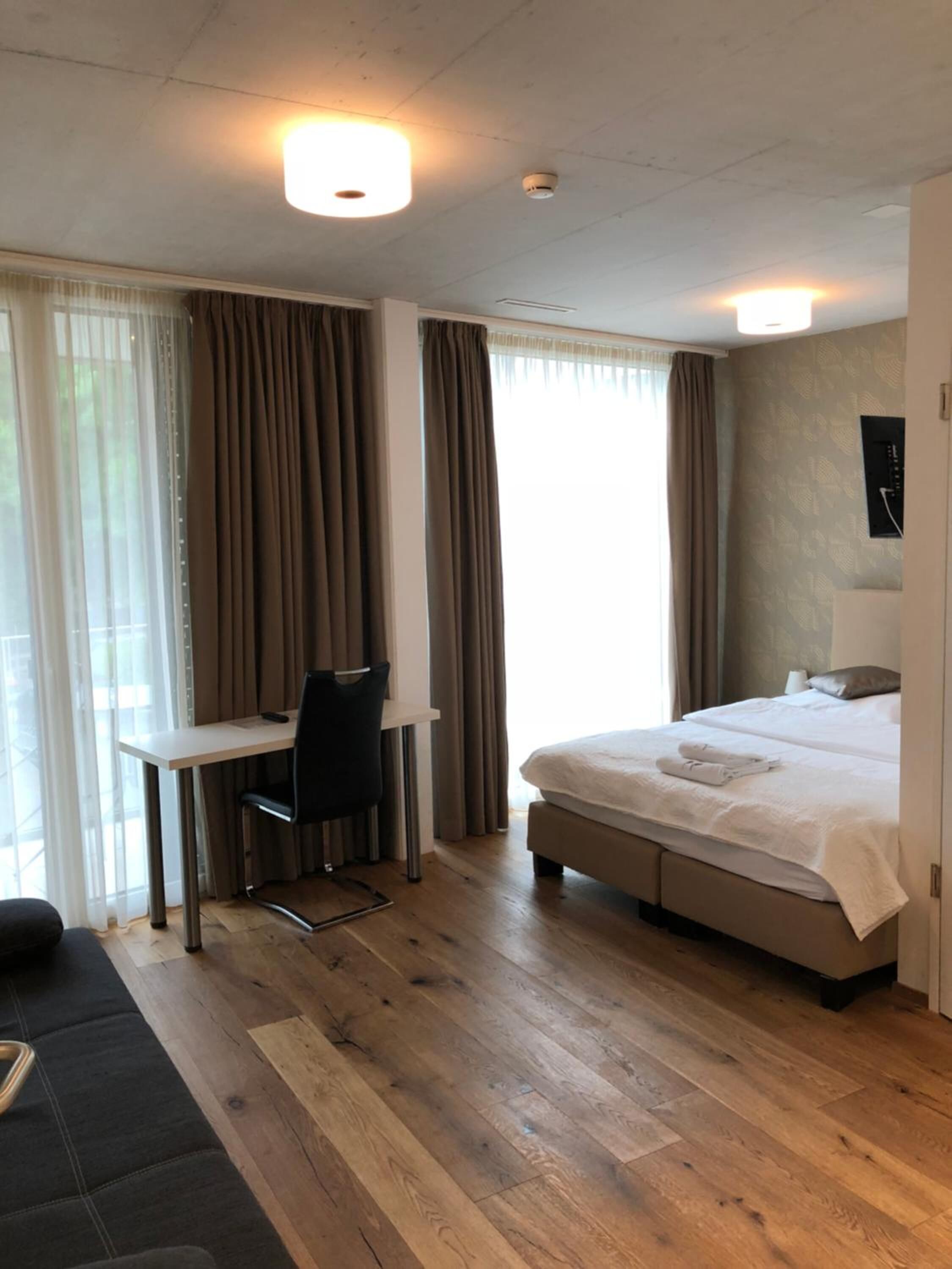 Une chambre d’hôtel moderne avec un plancher en bois, un lit avec du linge de maison blanc, un bureau avec une chaise et de grandes fenêtres avec des rideaux.