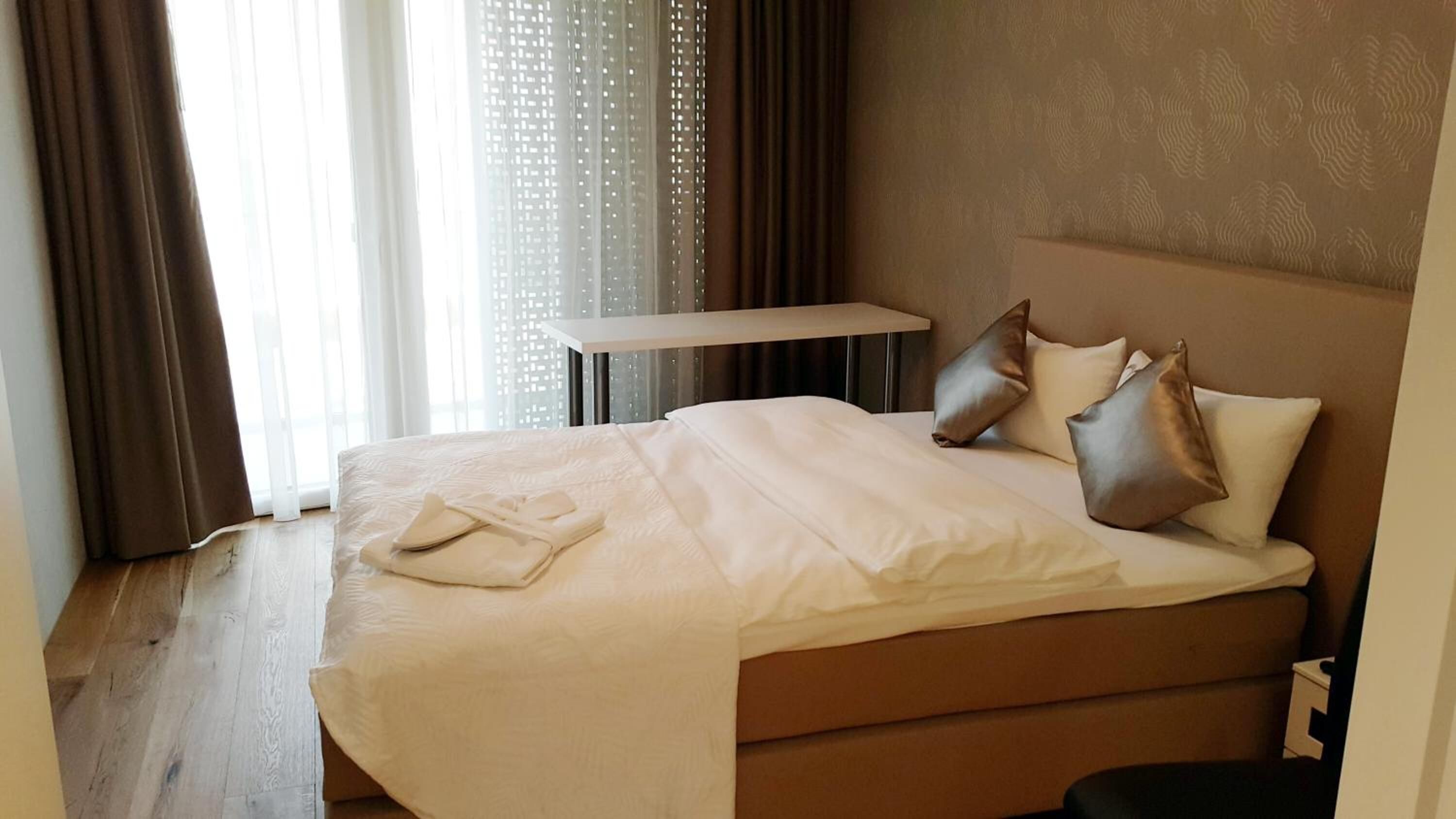 A neatly made bed with a white comforter and pillows, a small bedside table, and a view of a window with curtains.