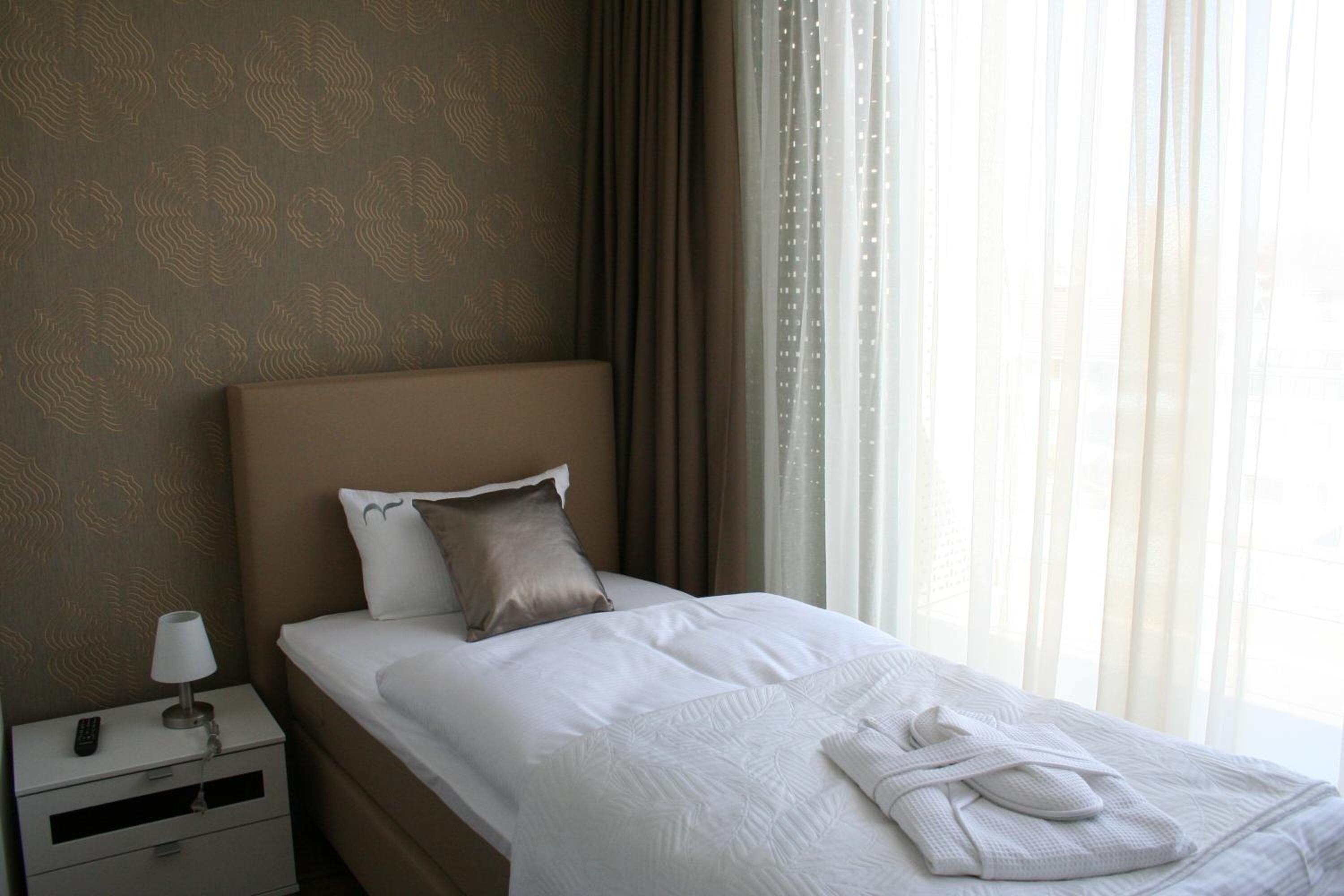 A neatly made bed with white linens and a folded towel, a bedside lamp, and a remote control on a nightstand.