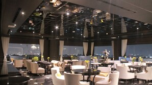 A modern restaurant with white chairs and tables, large windows, and a city view. - Lafayette Boutique Hotel Yogyakarta (Depok)