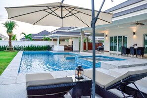 A swimming pool with lounge chairs and a large umbrella, surrounded by a modern house with a patio area. - Tulip House Pool Villa Hua Hin (Cha-am)