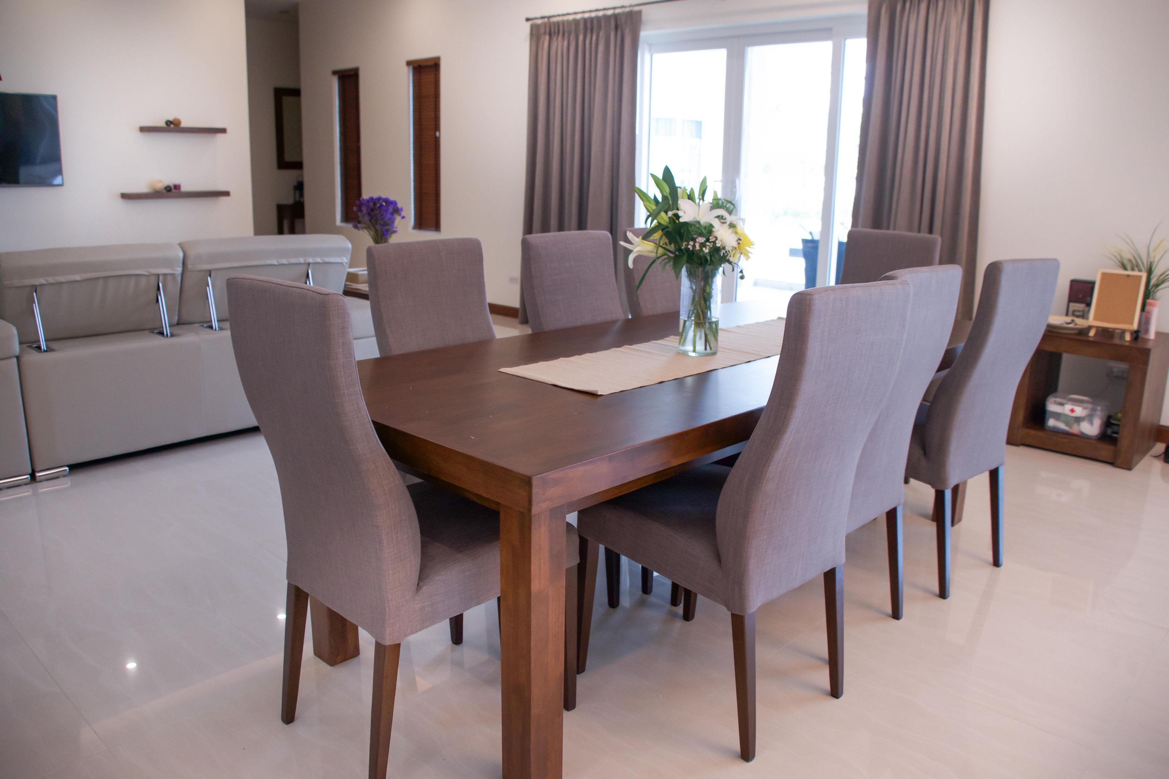 A modern dining area with a wooden table and six grey upholstered chairs. A vase with flowers is placed on the table.
