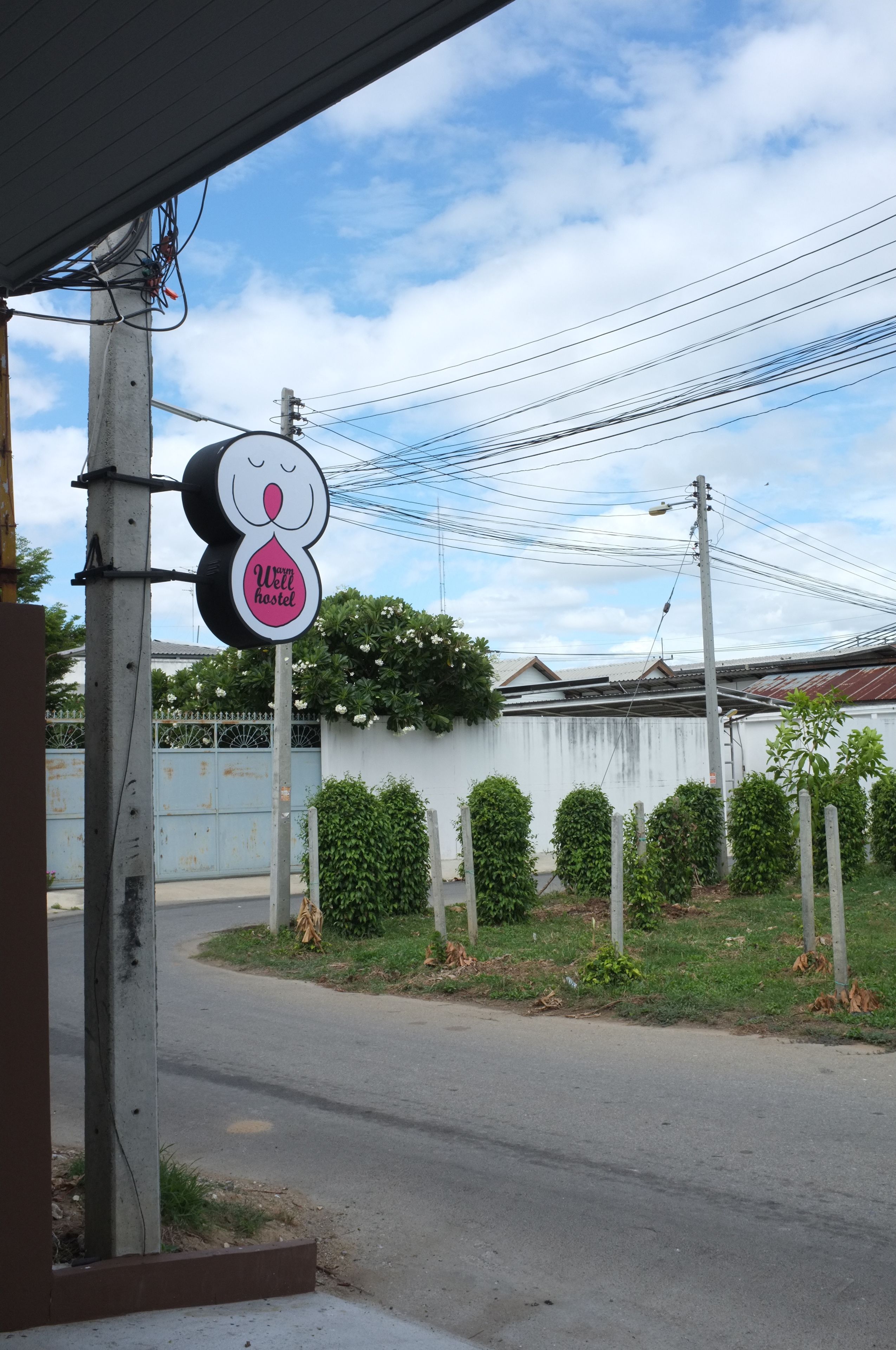 Señal de un hostal con un rostro y el texto 'Well Hostel' sobre un cielo despejado, líneas eléctricas y un área cercada con follaje.