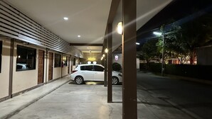 Un espace de stationnement couvert où est garée une voiture blanche, un bâtiment doté d’un toit en pente et un lampadaire visible au loin.