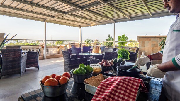 An outdoor patio with wicker furniture, a table with fruits and vegetables, and a chef preparing food. - Le Roi Jammu (Jammu)