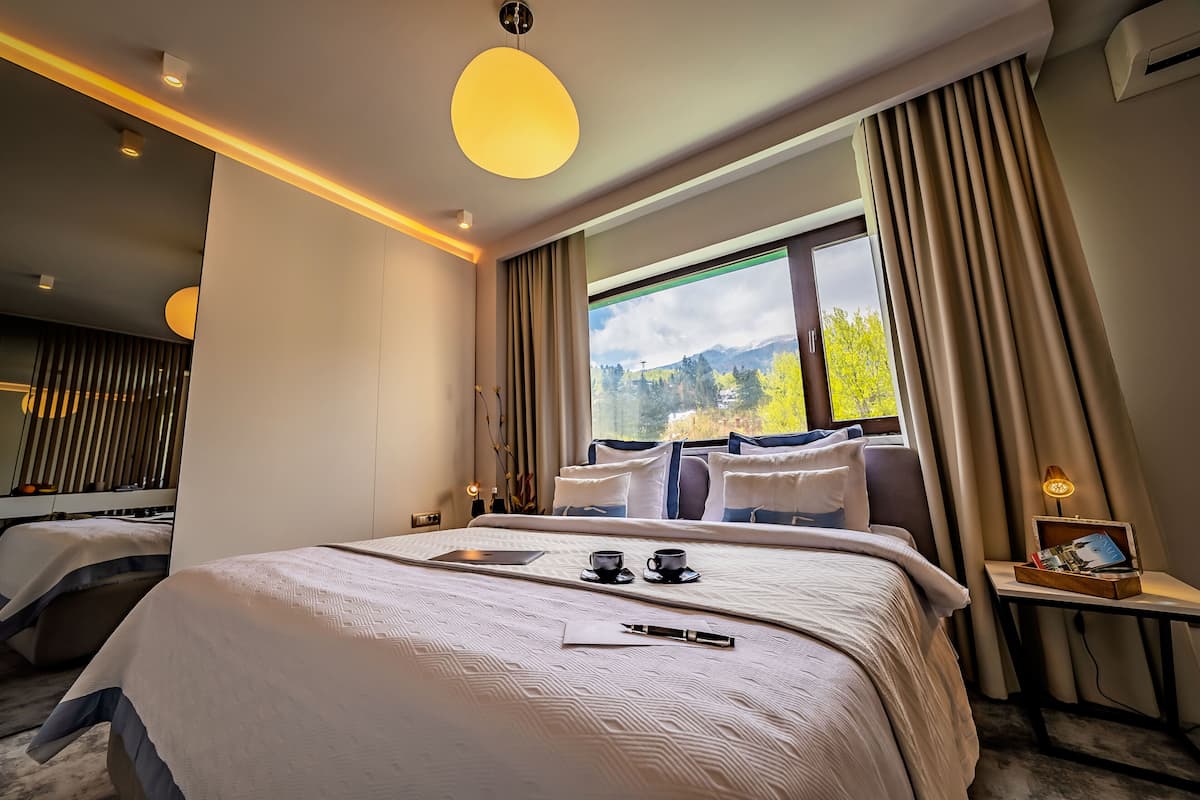 A modern hotel room with a large bed, a bedside table with a lamp, and a view of mountains through the window.