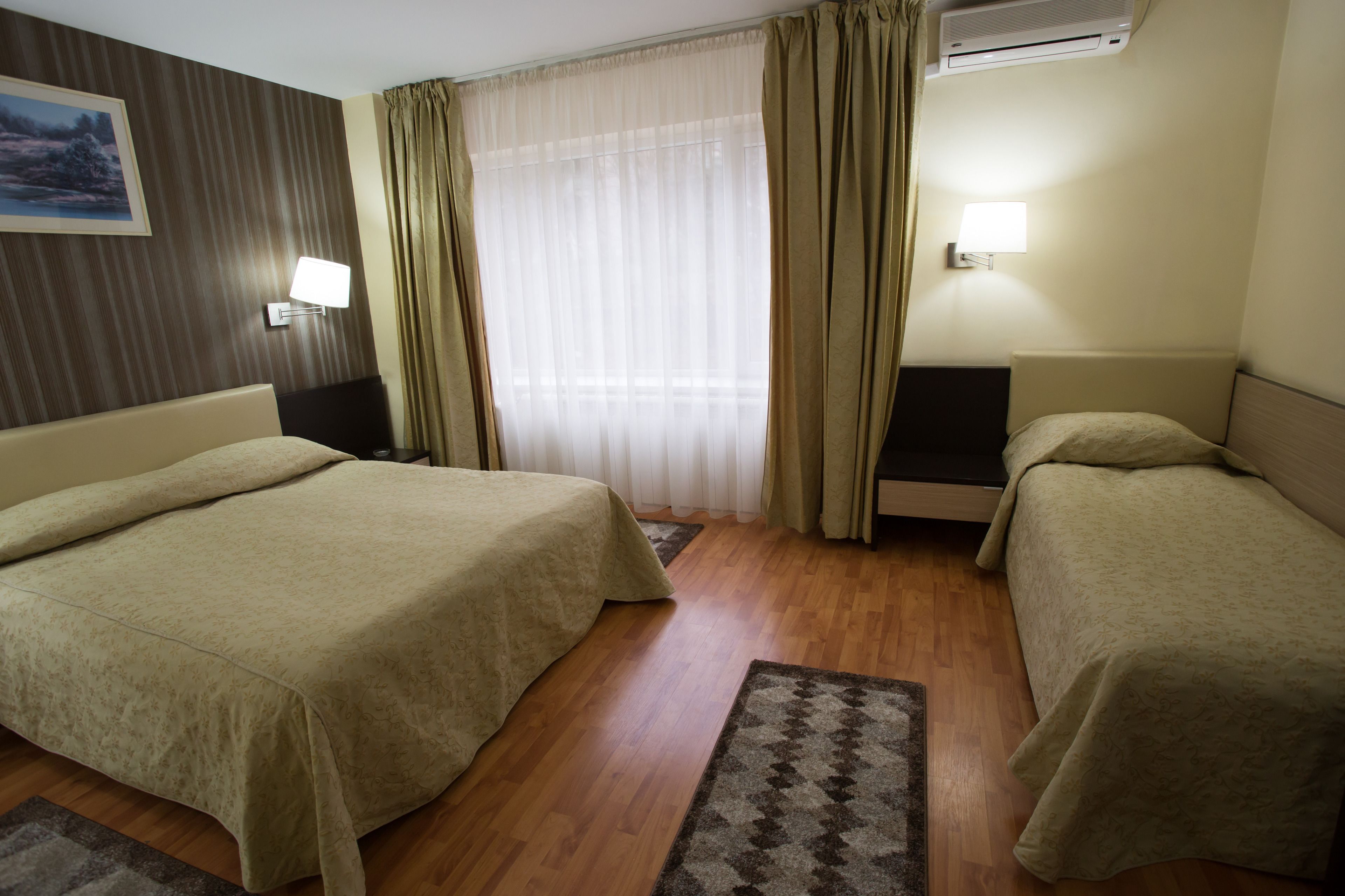 A hotel room with two beds, a wooden floor, a window with sheer curtains, a wall-mounted air conditioner, and a painting on the wall.