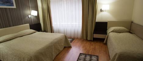 A hotel room with two beds, a wooden floor, a window with sheer curtains, a wall-mounted air conditioner, and a painting on the wall.