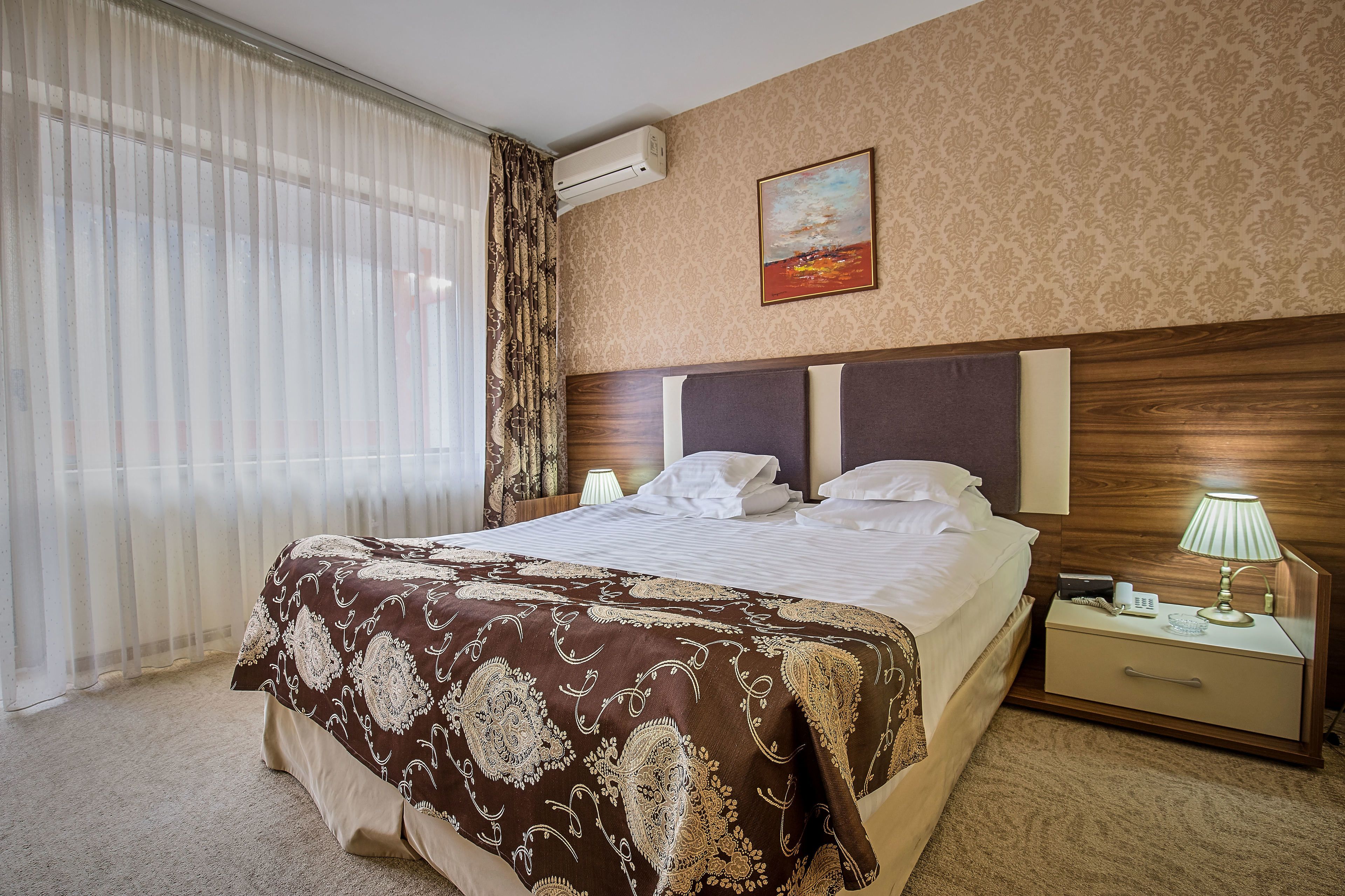 Habitación de hotel con una cama grande, una mesita de noche con lámpara, aire acondicionado en la pared y una ventana con cortinas traslúcidas.