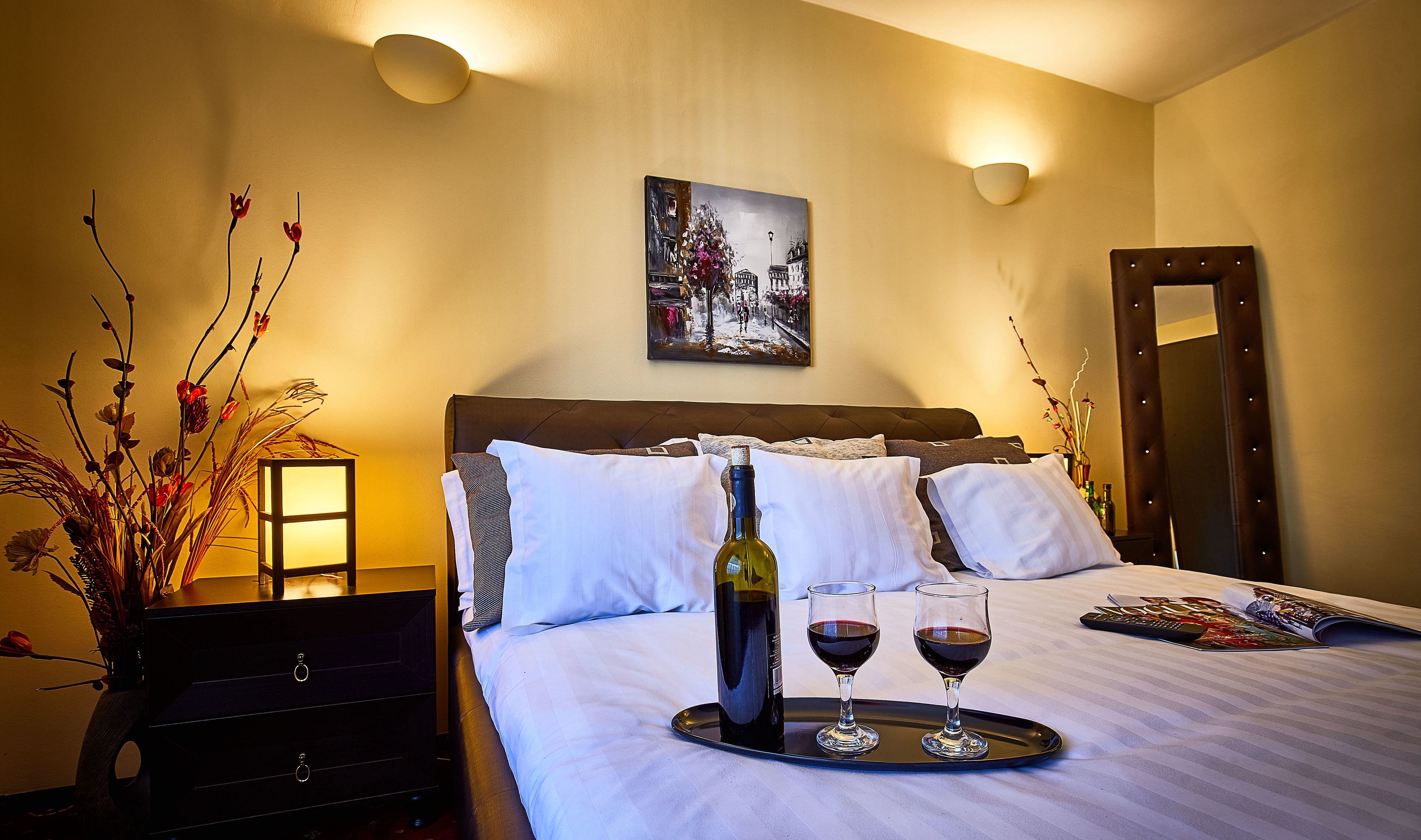 Habitación de hotel con cama, mesita de noche con una botella de vino y dos vasos, un cuadro en la pared y un espejo.