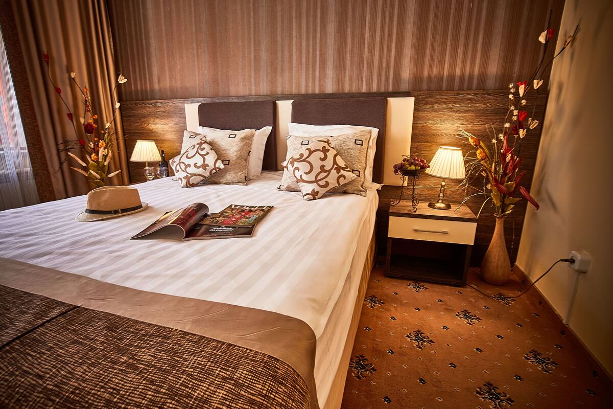 A neatly made bed with a magazine, a hat, and decorative pillows. There are two bedside tables with lamps and a vase of flowers. The room has a wooden headboard and patterned carpet.