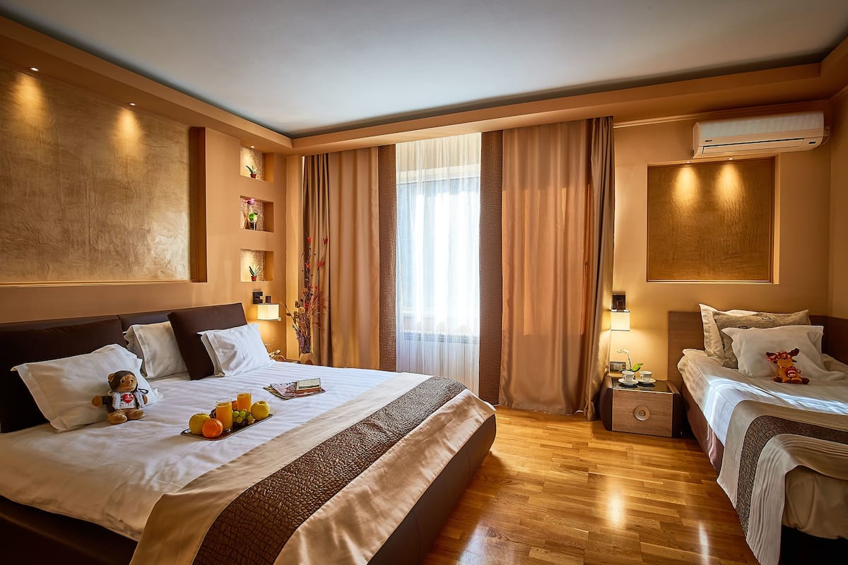 A hotel room with a large bed, a smaller bed, an air conditioning unit, a nightstand, and a window with curtains.
