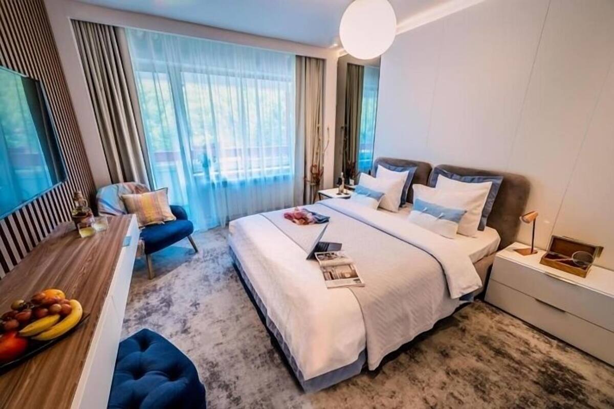 A modern hotel room with a large bed, a desk with fruit, a chair, and a view of the outdoors.