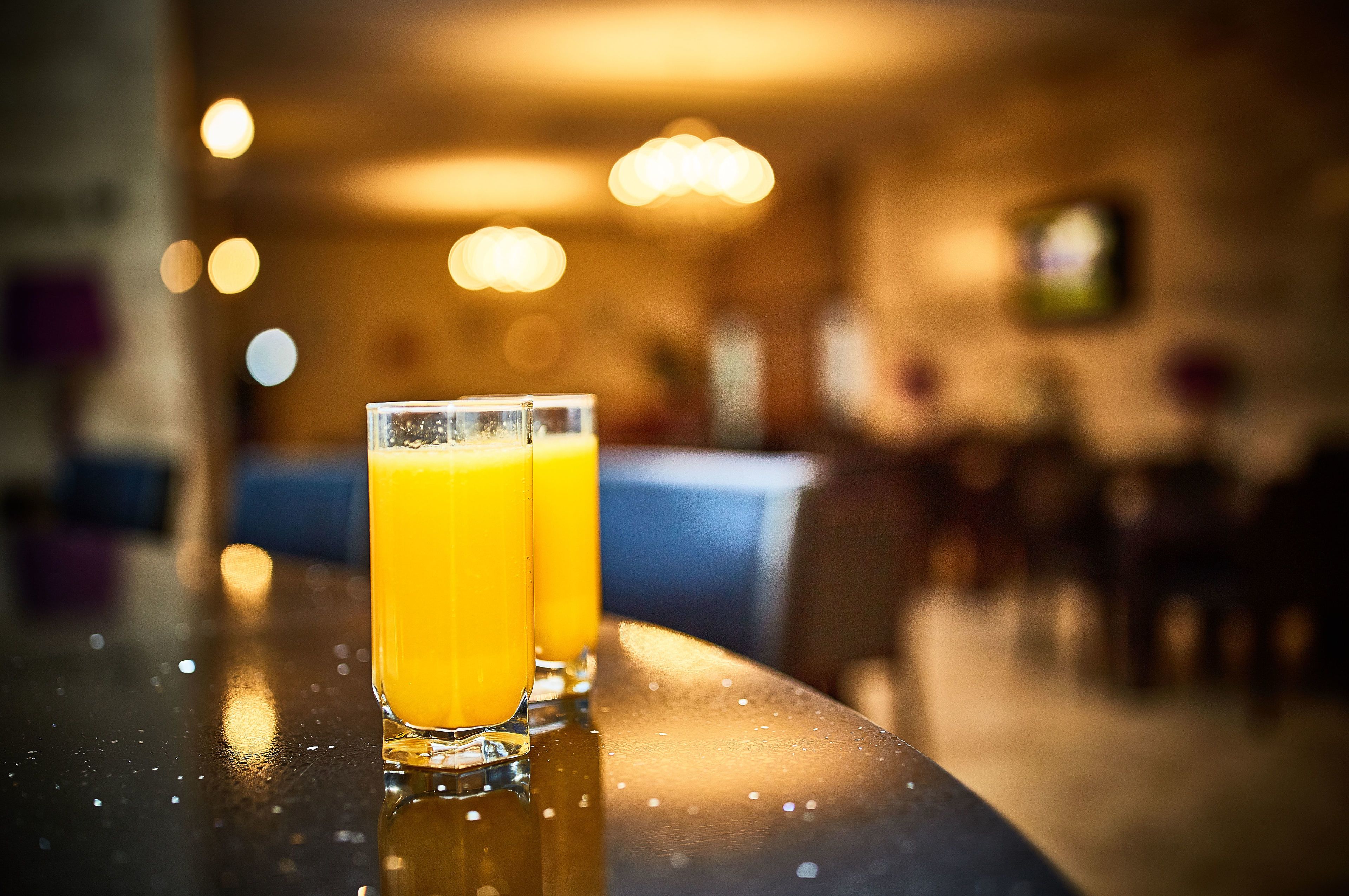 Two glasses of orange juice on a reflective surface in a dimly lit room with blurred background lights and furniture.