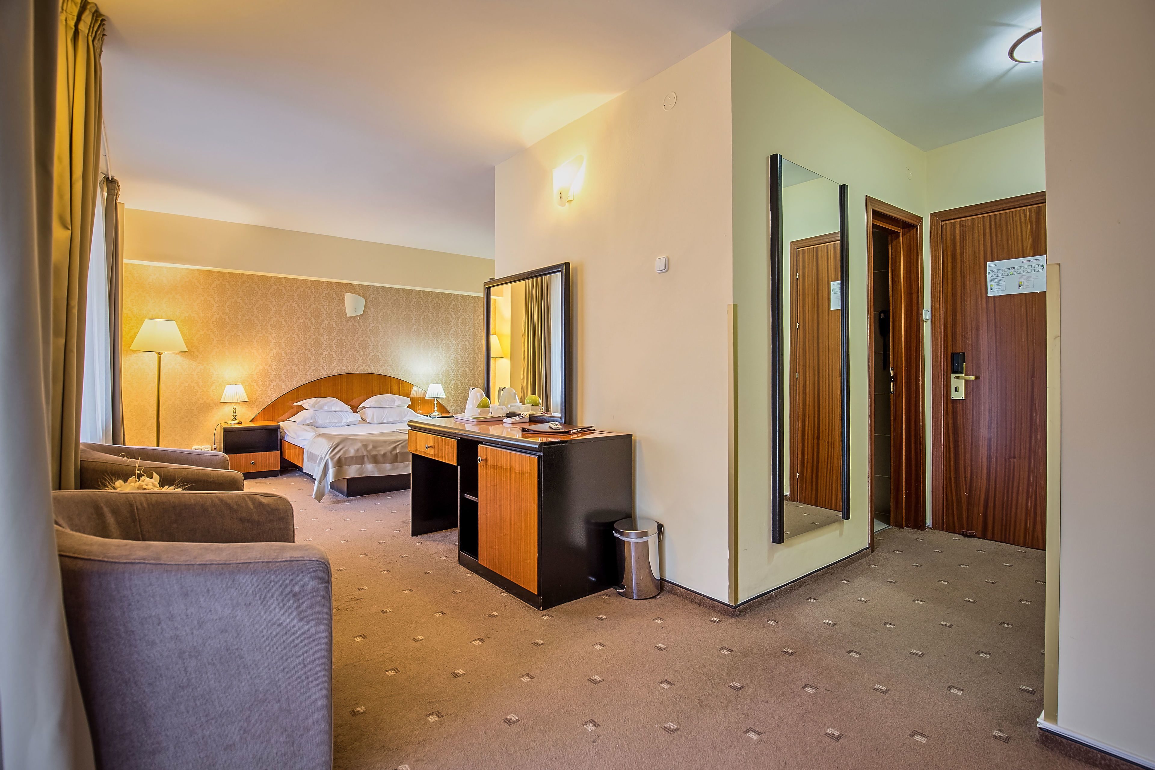 A hotel room with a bed, a desk, a chair, a lamp, a trash bin, and a mirror.
