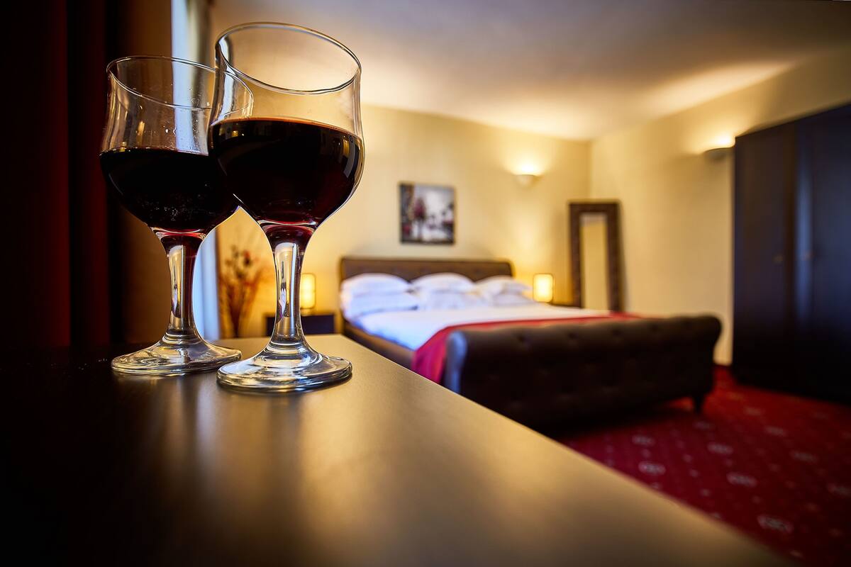 Two wine glasses on a table with a bed and a nightstand in the background.