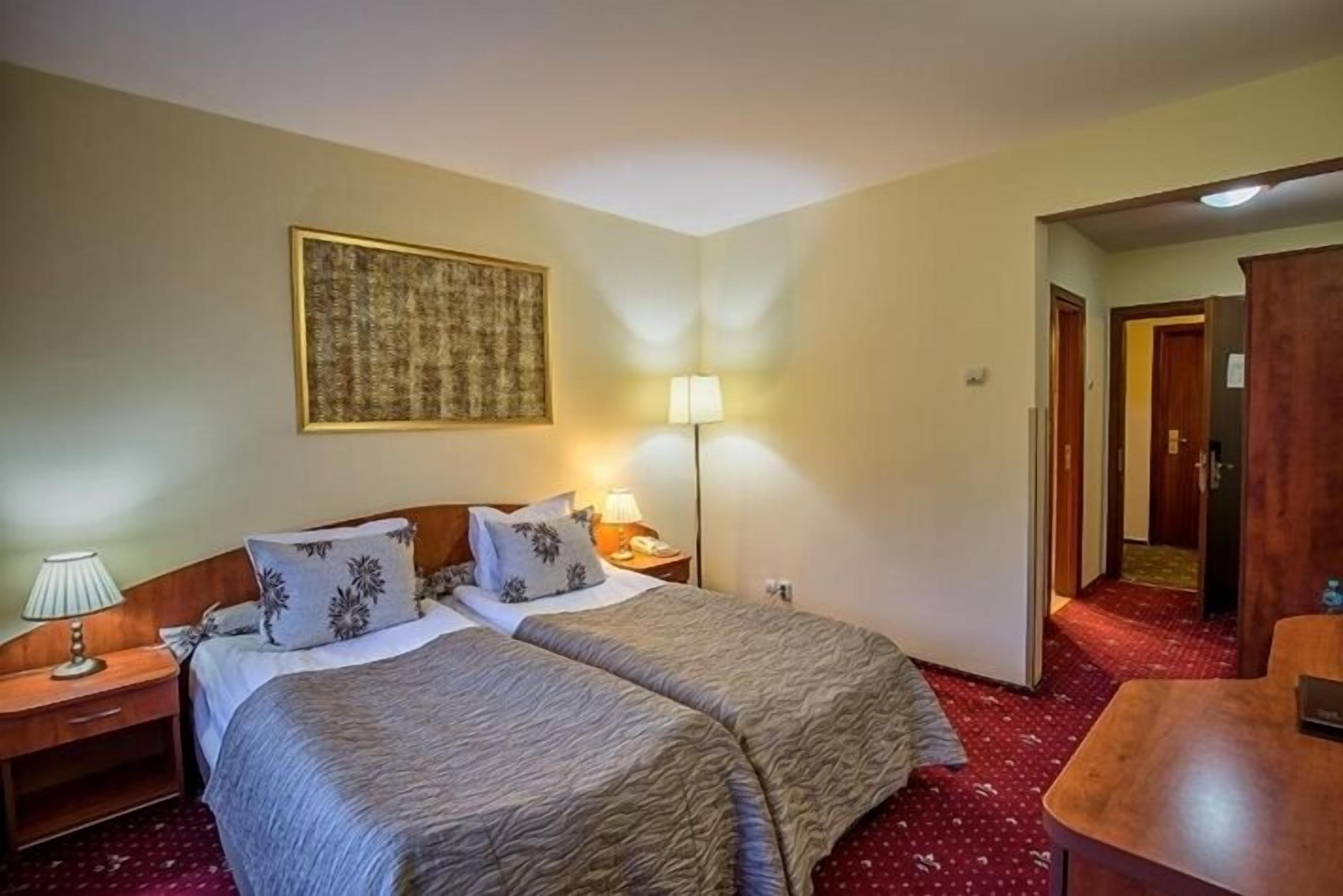 A hotel room with two beds, a nightstand with a lamp, a wall-mounted artwork, and a red carpeted floor.