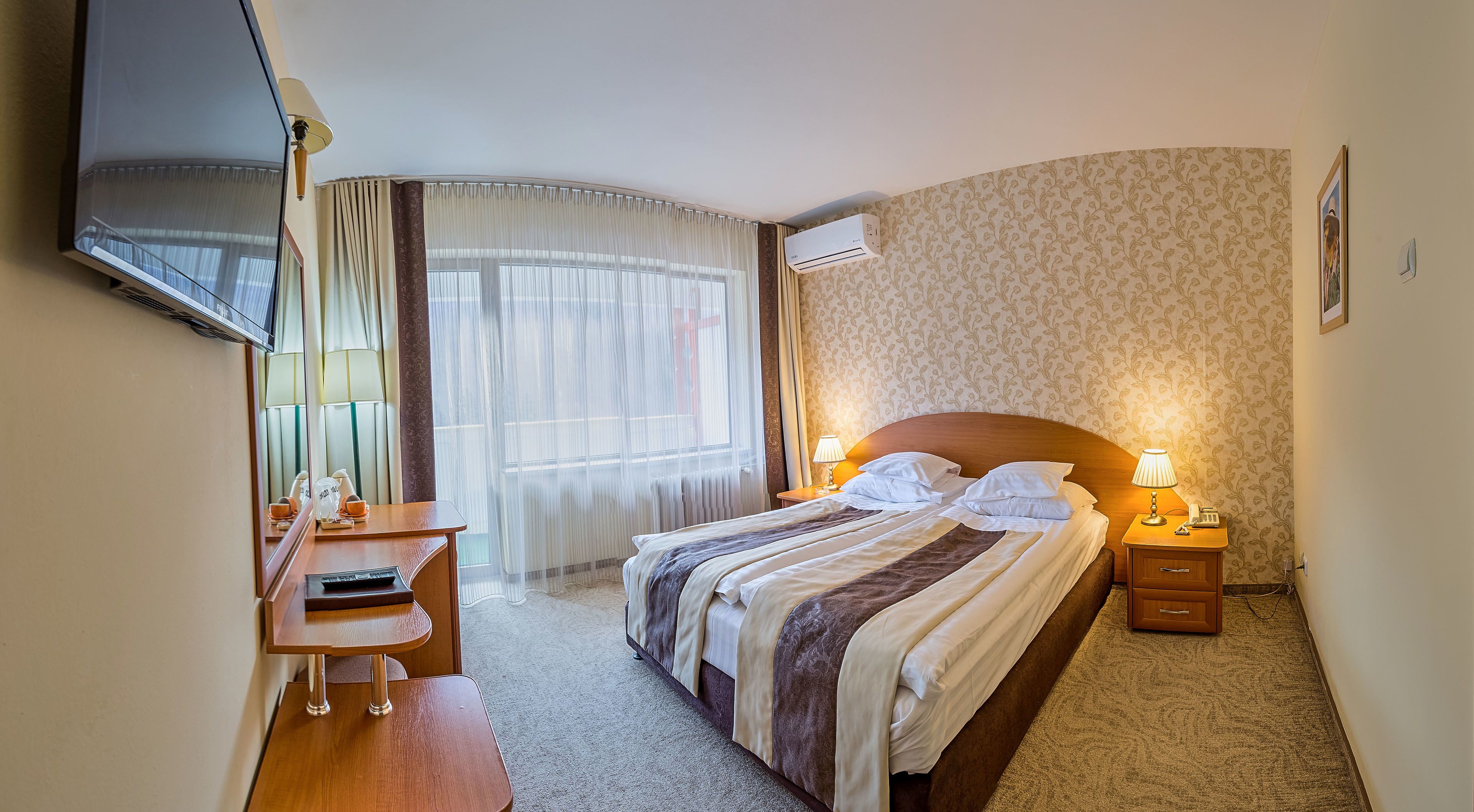 A hotel room with a bed, bedside tables, a desk, a chair, a TV, and an air conditioning unit.