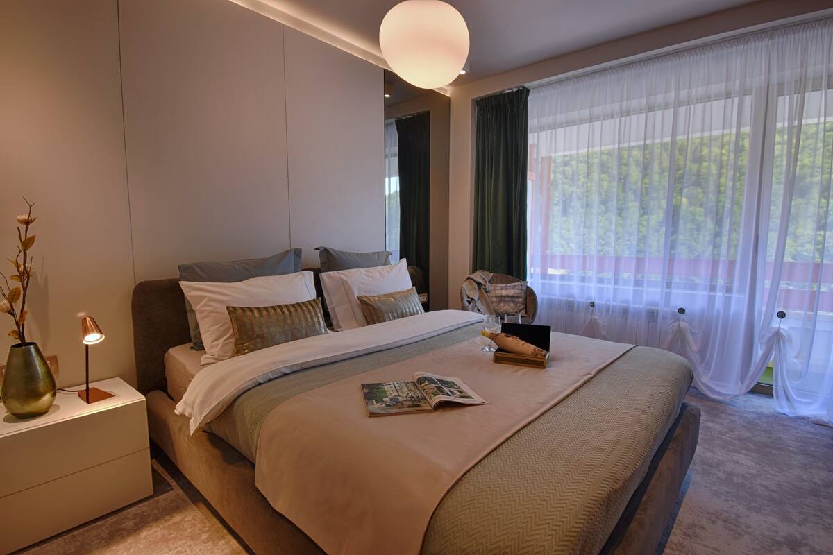 A modern bedroom with a large bed, a nightstand with a lamp, and a view of greenery through sheer curtains.