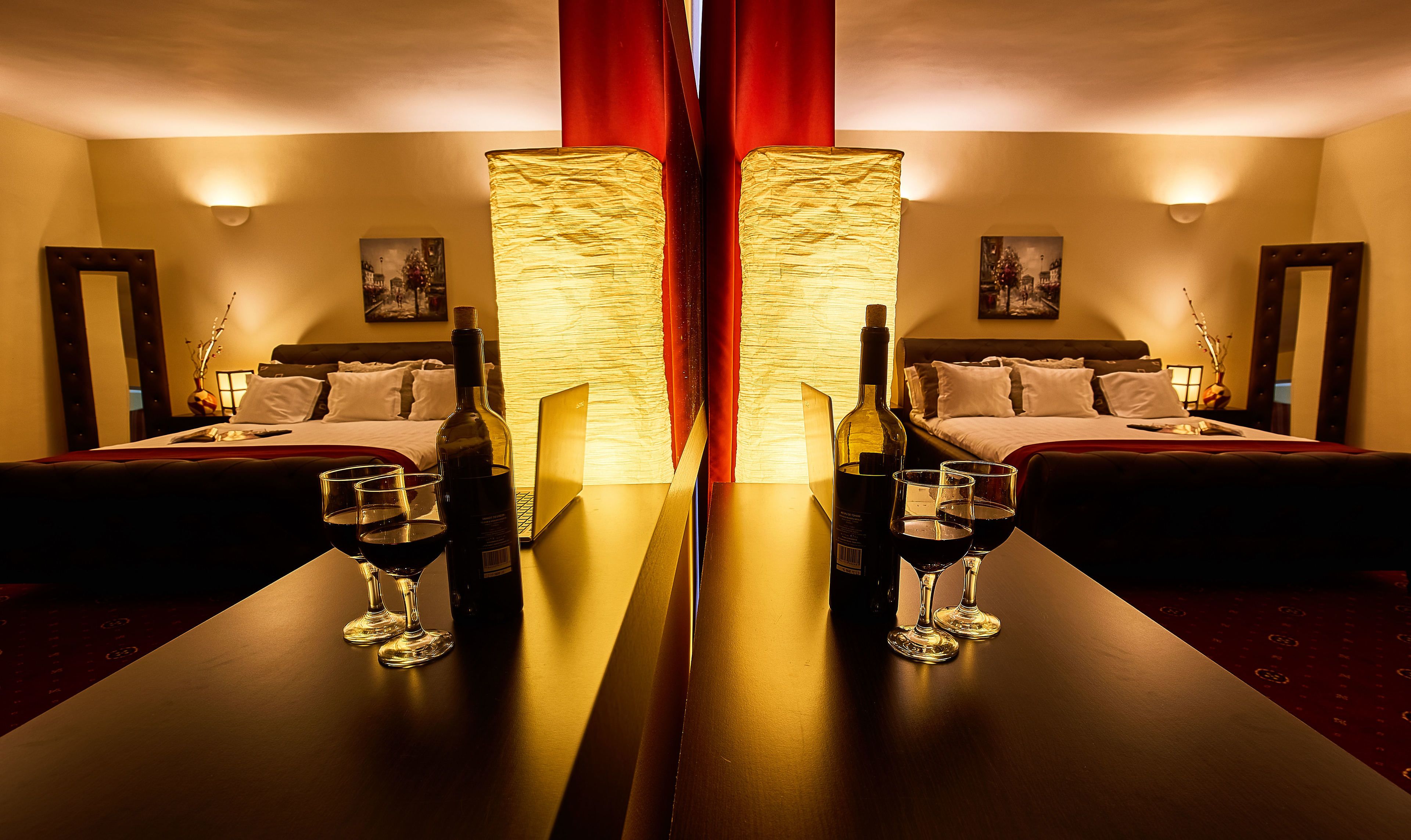 A mirrored room with a bed, a table with wine glasses and bottles, and a lamp.