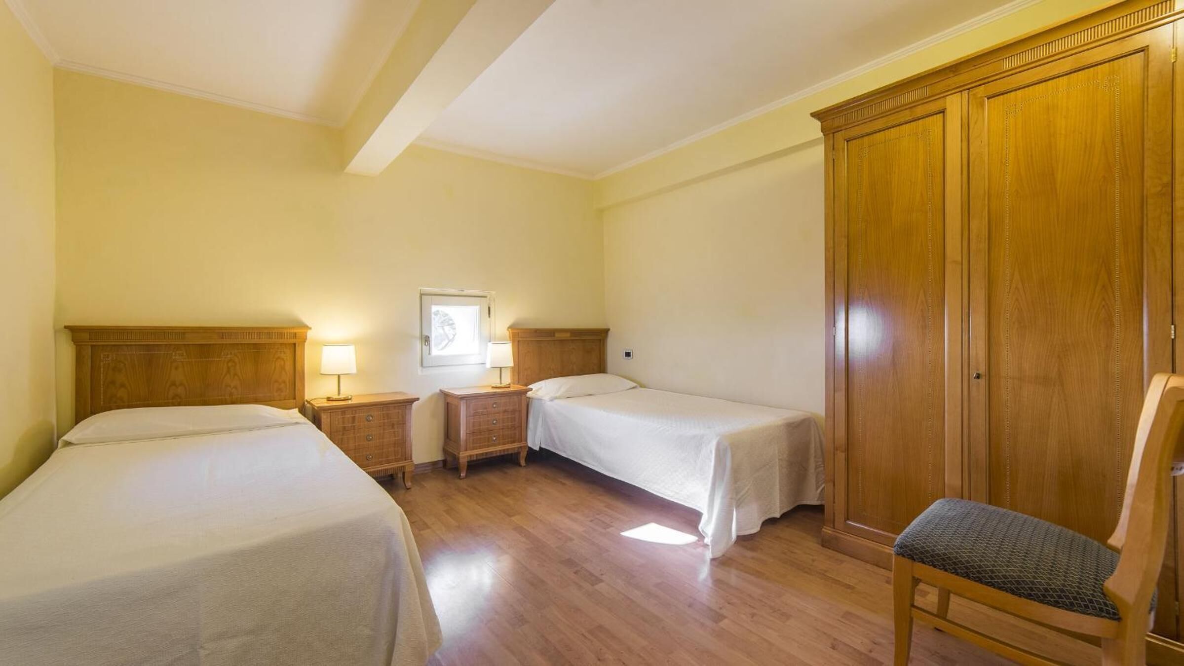 A hotel room with two beds, wooden headboards, a wardrobe, a chair, and bedside tables with lamps.