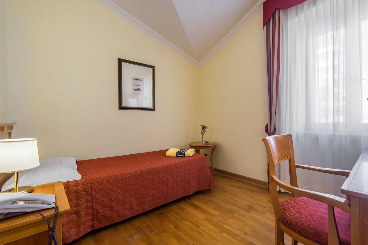 Ein Hotelzimmer mit einem Einzelbett, einem Schreibtisch mit Lampe, einem Sessel, einem Fernseher und einem gerahmten Bild an der Wand.
