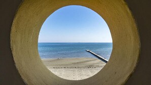Vista a través de un ventanal circular de una playa con un muelle que se adentra en el agua.