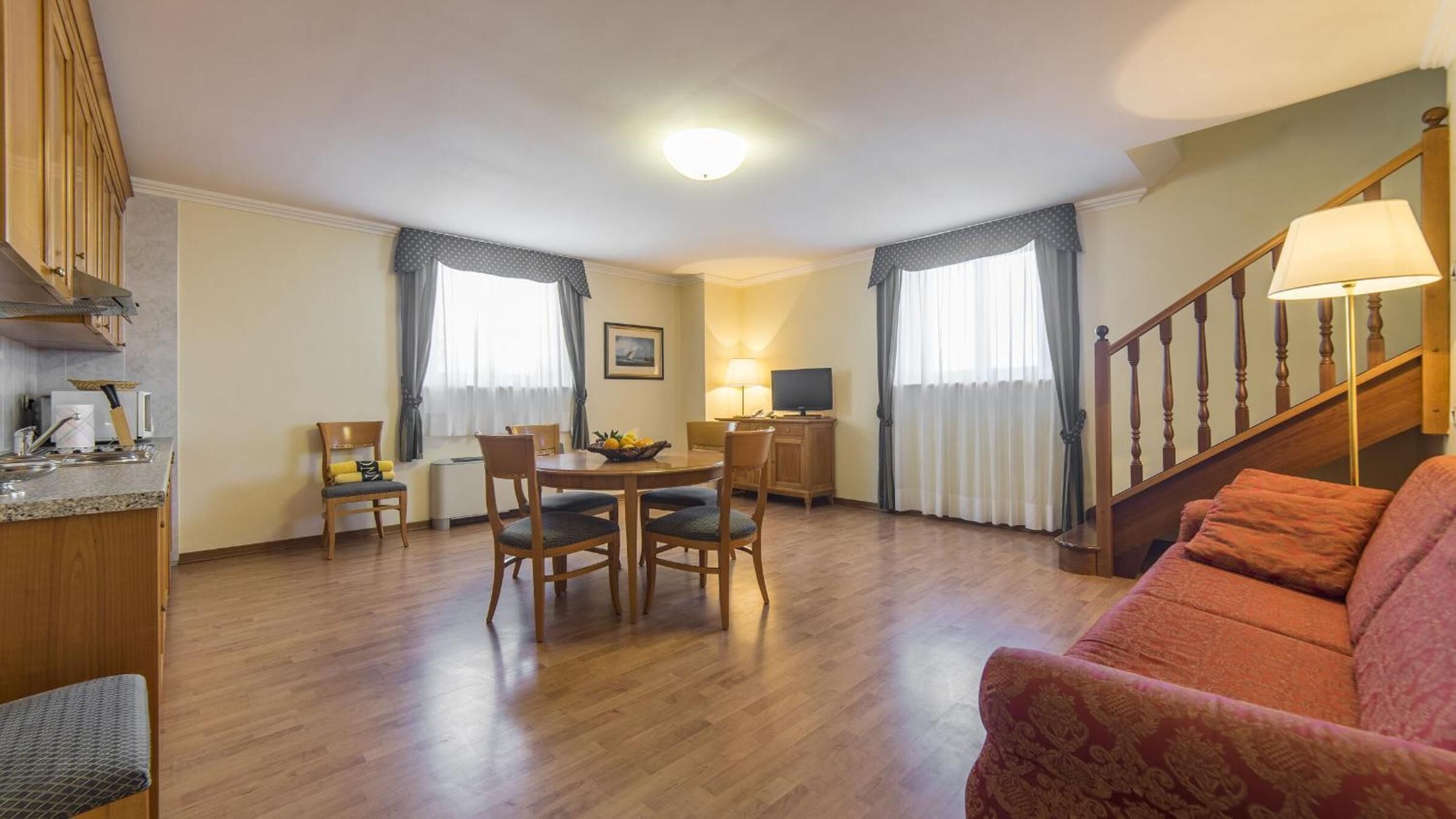 A well-lit living room with a dining area, wooden furniture, a staircase, and a kitchen area with a microwave.