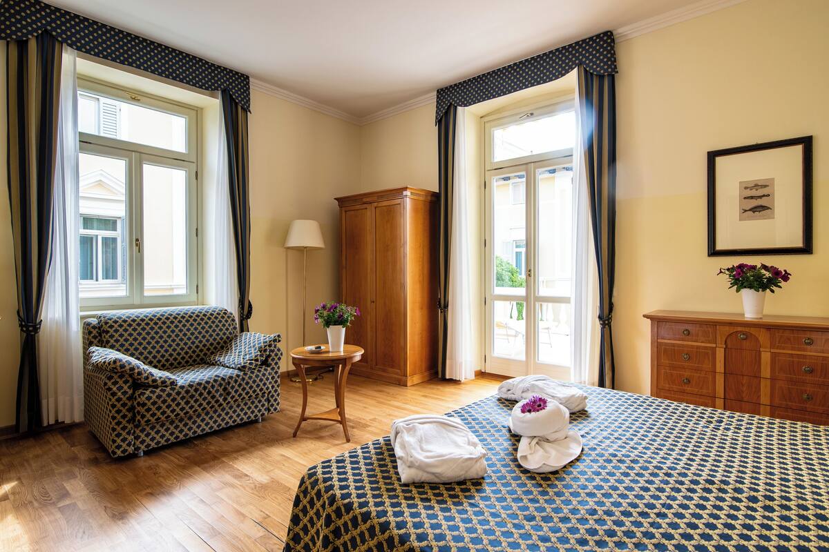 Ein Hotelzimmer mit einer gestreiften Couch, einem Bett mit blau-weiß karierter Bettdecke, einem Holzschrank und einem kleinen Tisch mit Blumen in einer Vase.