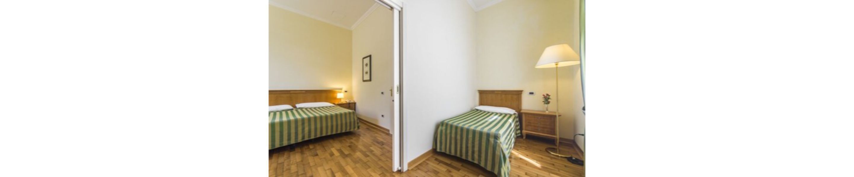 A hotel room with two beds, wooden flooring, a nightstand with a lamp, and a door.