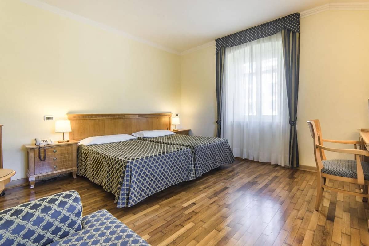 Ein Hotelzimmer mit einem hölzernen Bett, einem Schreibtisch mit Computer, einem Stuhl und einem Nachttisch mit Lampe.