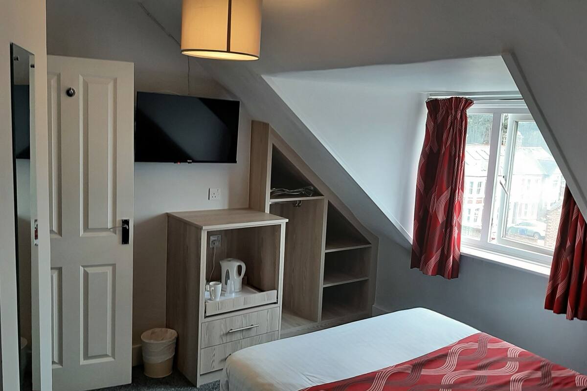 A bedroom with a bed, a TV mounted on the wall, a wooden cabinet, a trash bin, and a window with curtains.