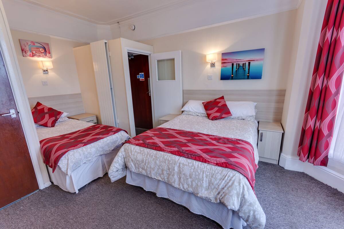 A hotel room with two beds, each with red and white bedding, a nightstand, a wall-mounted lamp, and a painting above the bed.