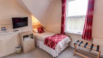 A bedroom with a bed, a TV, a radiator, a window with curtains, and a small cabinet with items.