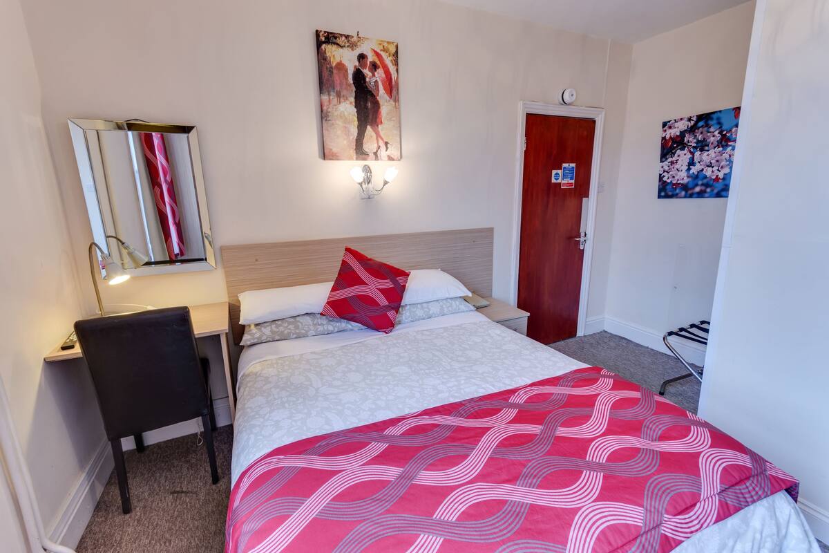 A hotel room with a bed, a desk, a chair, a mirror, and a door.