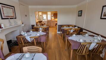 A dining area with multiple tables set for meals, wooden chairs, and a kitchen visible in the background.