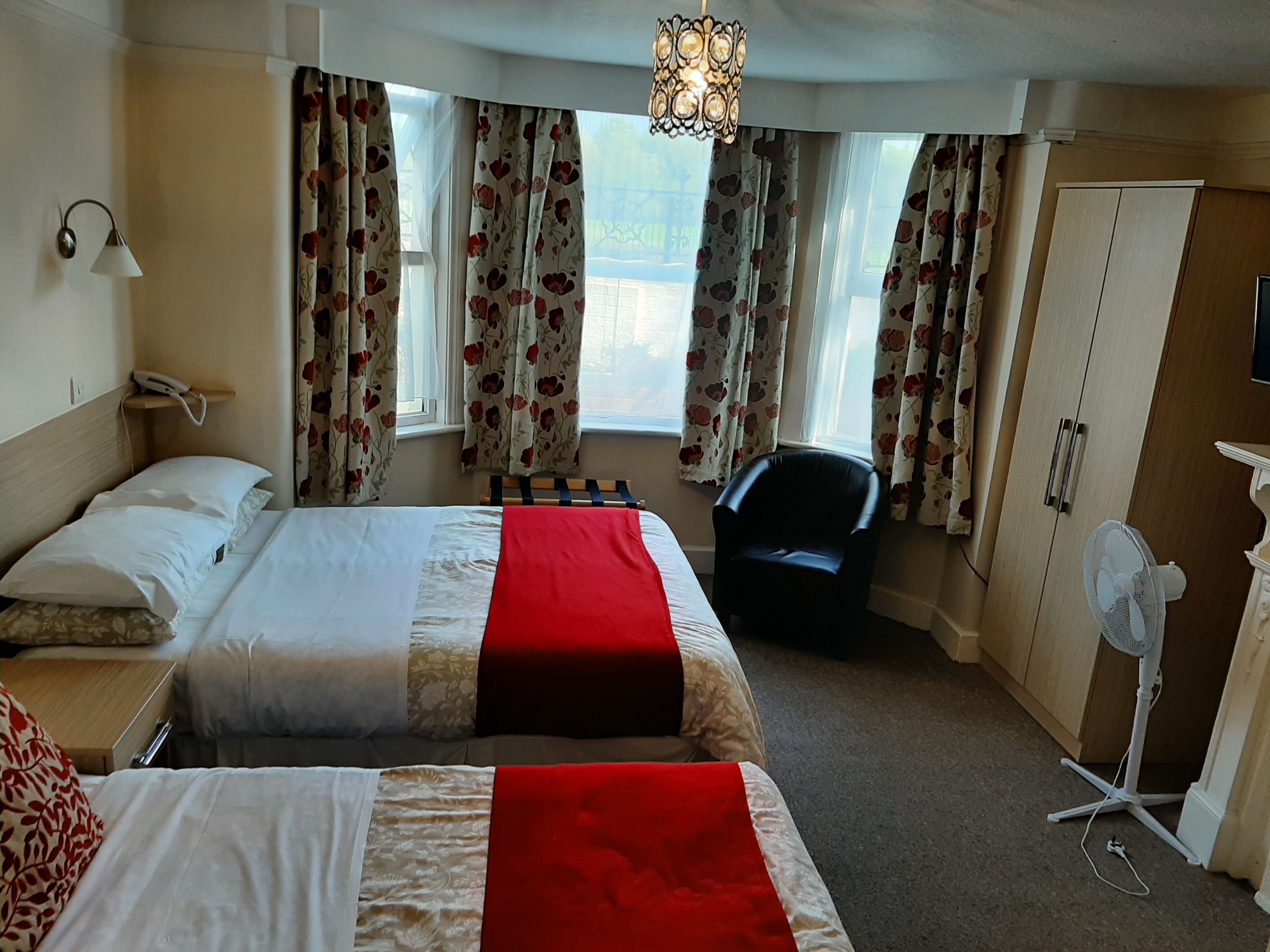 A hotel room with two beds, a desk, a chair, a wardrobe, and a fan.