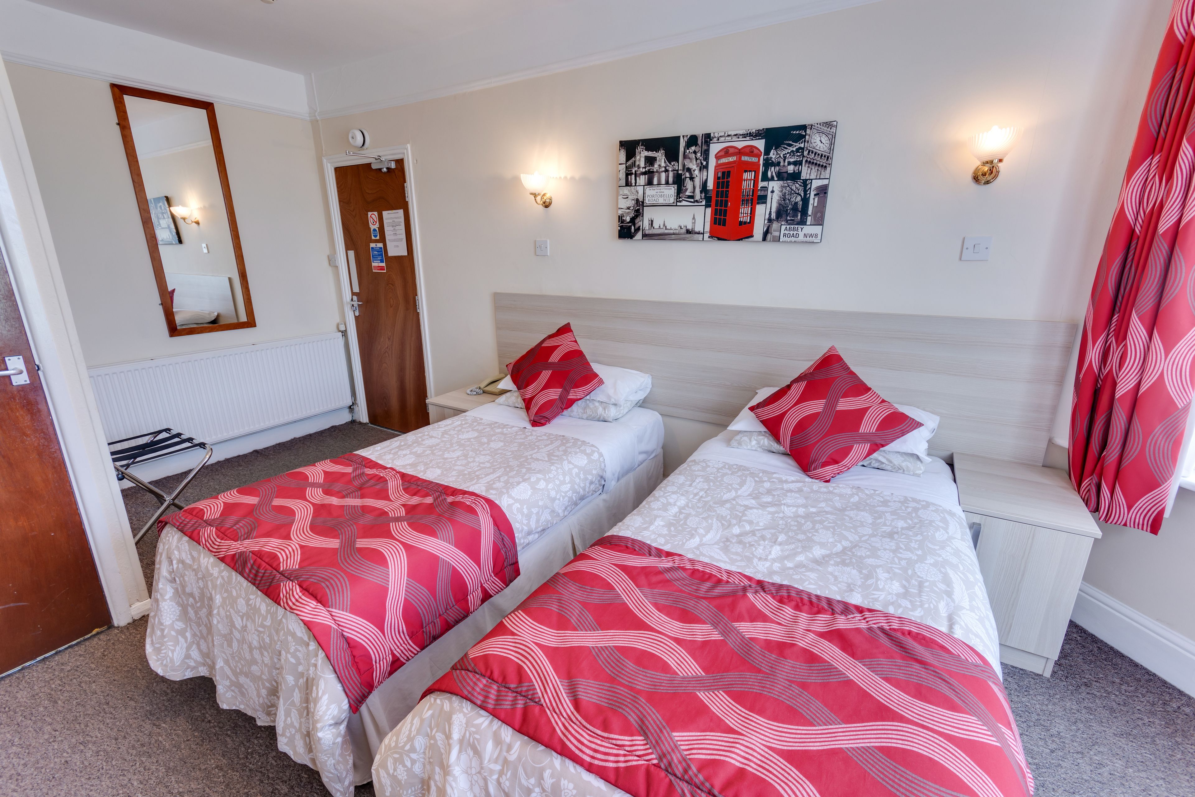 A hotel room with two beds, each with red and white bedding, a wooden headboard, a small bedside table, and a wall-mounted light fixture.