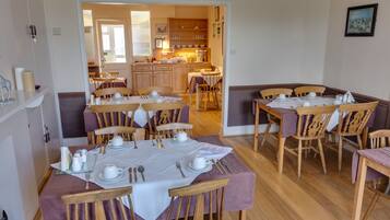 A dining area with multiple tables set for meals, wooden chairs, and a kitchen in the background.