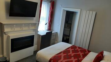 A hotel room with a fireplace, a television mounted above it, a bed with red and white bedding, and a small desk with a lamp.