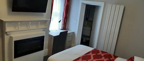 A hotel room with a fireplace, a television mounted above it, a bed with red and white bedding, and a small desk with a lamp.