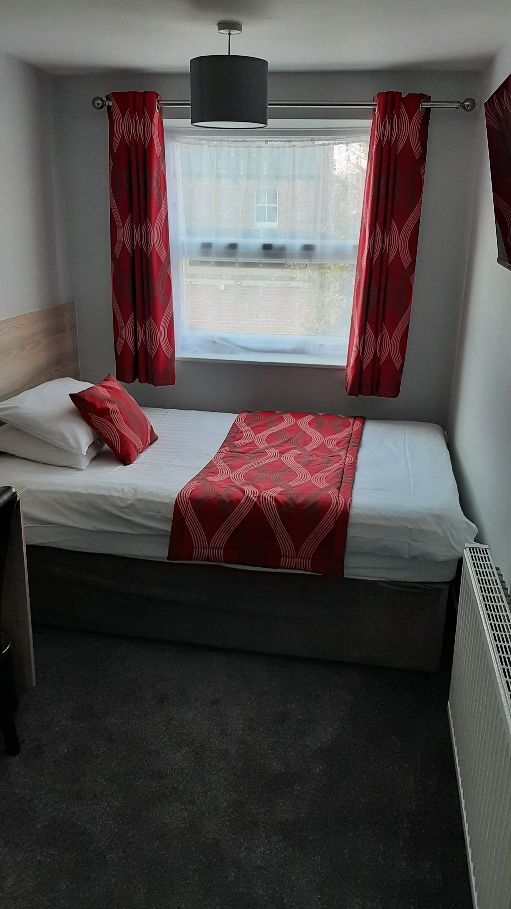 Ein Einzelbett mit einer rot gemusterten Decke, weißen Kissen und roten Vorhängen.