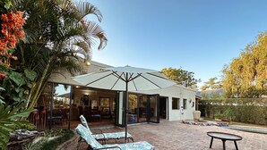 An outdoor patio with a brick floor, two lounge chairs, a small table, and a large umbrella. - Malcolm Lodge (Harare)