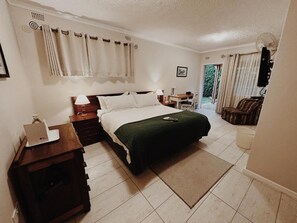 A bedroom with a bed, bedside tables, a desk, a chair, and a view of the outdoors. - Malcolm Lodge (Harare)