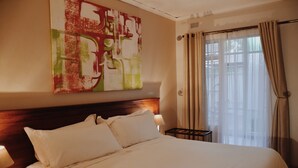 A hotel room with a large bed, a headboard, a nightstand with a lamp, and a window with curtains. - Malcolm Lodge (Harare)