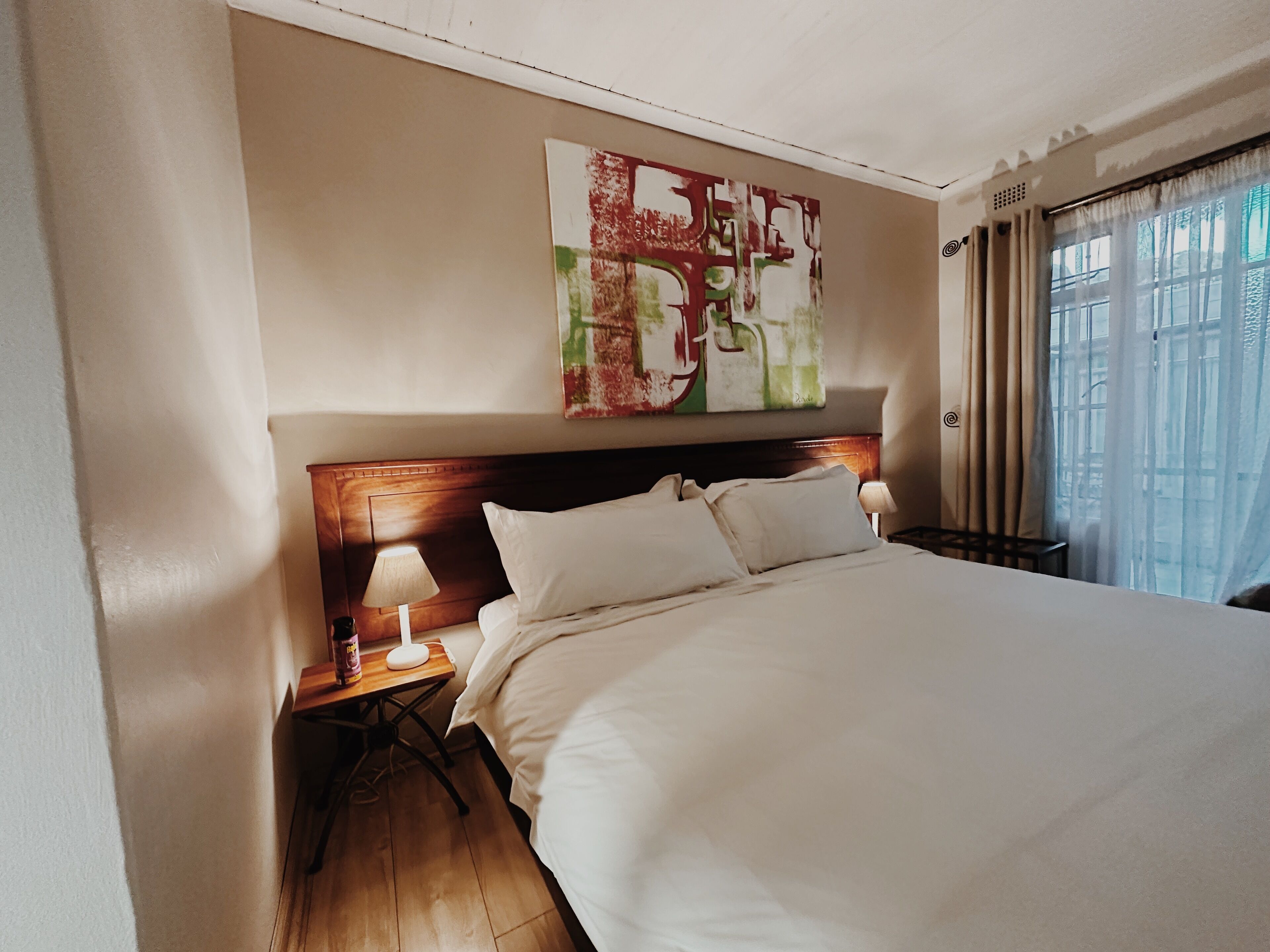 A hotel room with a large bed, a wooden headboard, a bedside table with a lamp, and a window with curtains.